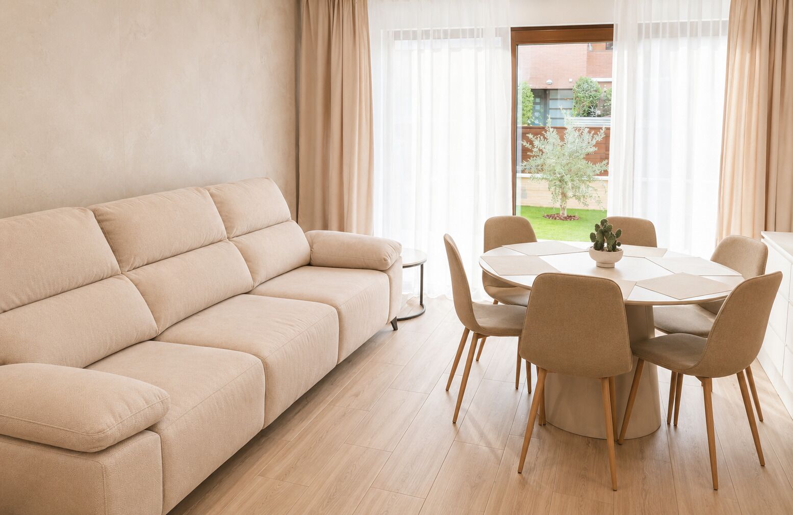 Una sala de estar con un sofá color beige, una mesa de comedor redonda con sillas y vista a un jardín a través de una ventana.