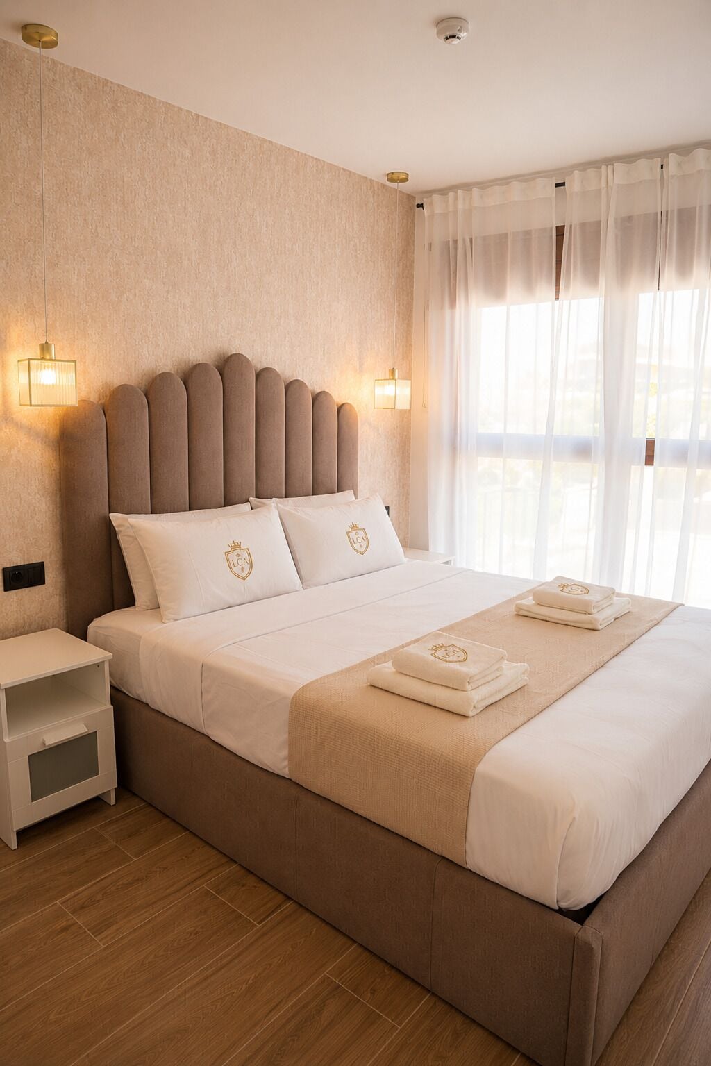 A hotel room with a large bed, bedside tables, and a window with sheer curtains.
