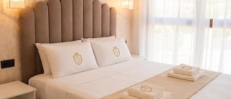 A hotel room with a large bed, bedside tables, and a window with sheer curtains.