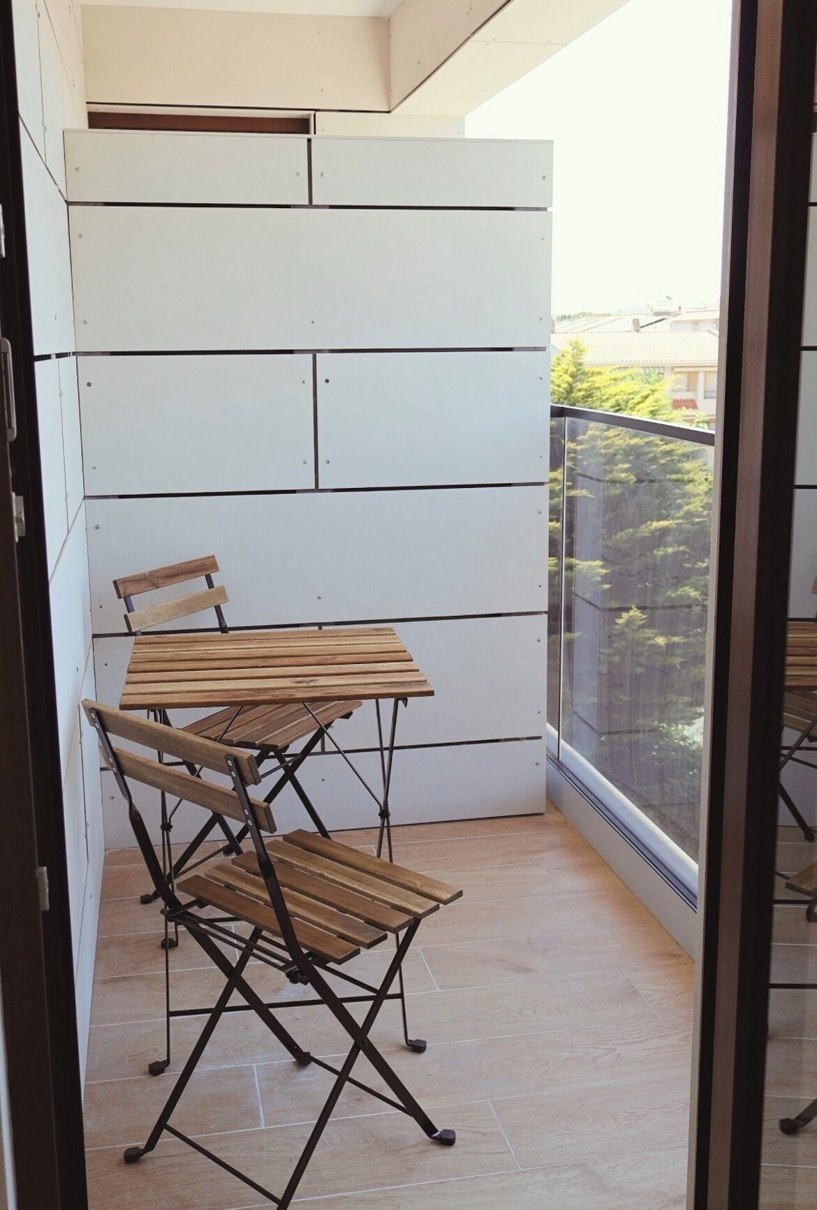 Un balcón con mesa y sillas de madera, una pared blanca y vistas a follaje verde.