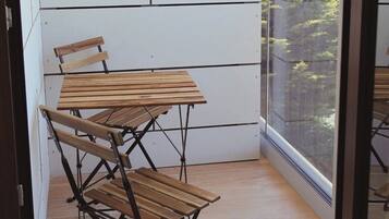 Un balcón con mesa y sillas de madera, una pared blanca y vistas a follaje verde.