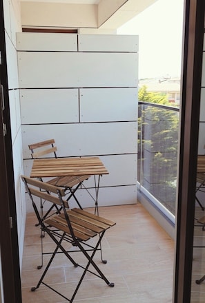 Un balcón con mesa y sillas de madera, una pared blanca y vistas a follaje verde.