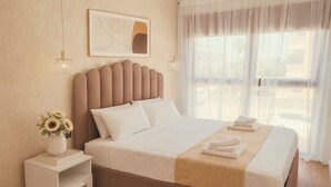 A hotel room with a bed, a bedside table with a vase of flowers, and a window with sheer curtains. - Luxury Cubelles Aparthotel (Cubelles)