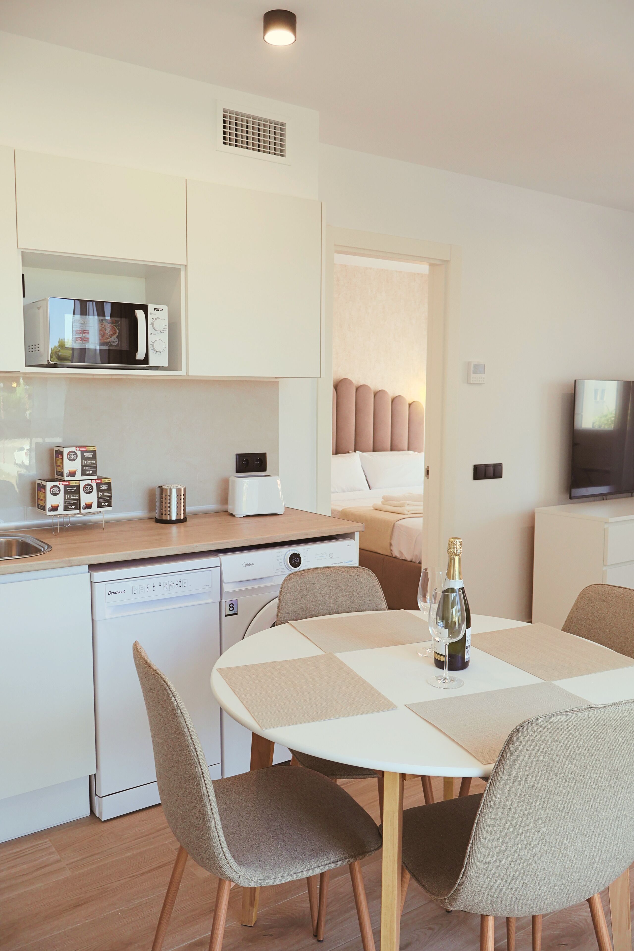 Cocina moderna equipada con microondas, tostadora y lavavajillas. Hay una zona de comedor con mesa y sillas, y una cama con cabecera al fondo.