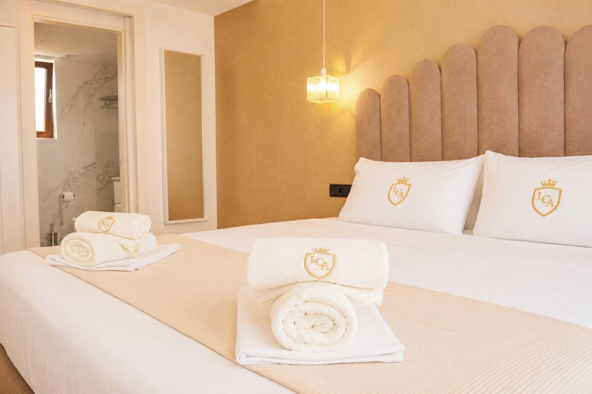 A hotel room with a bed, two pillows with a crest logo, a bedside lamp, and a towel with a crest logo on the bed.