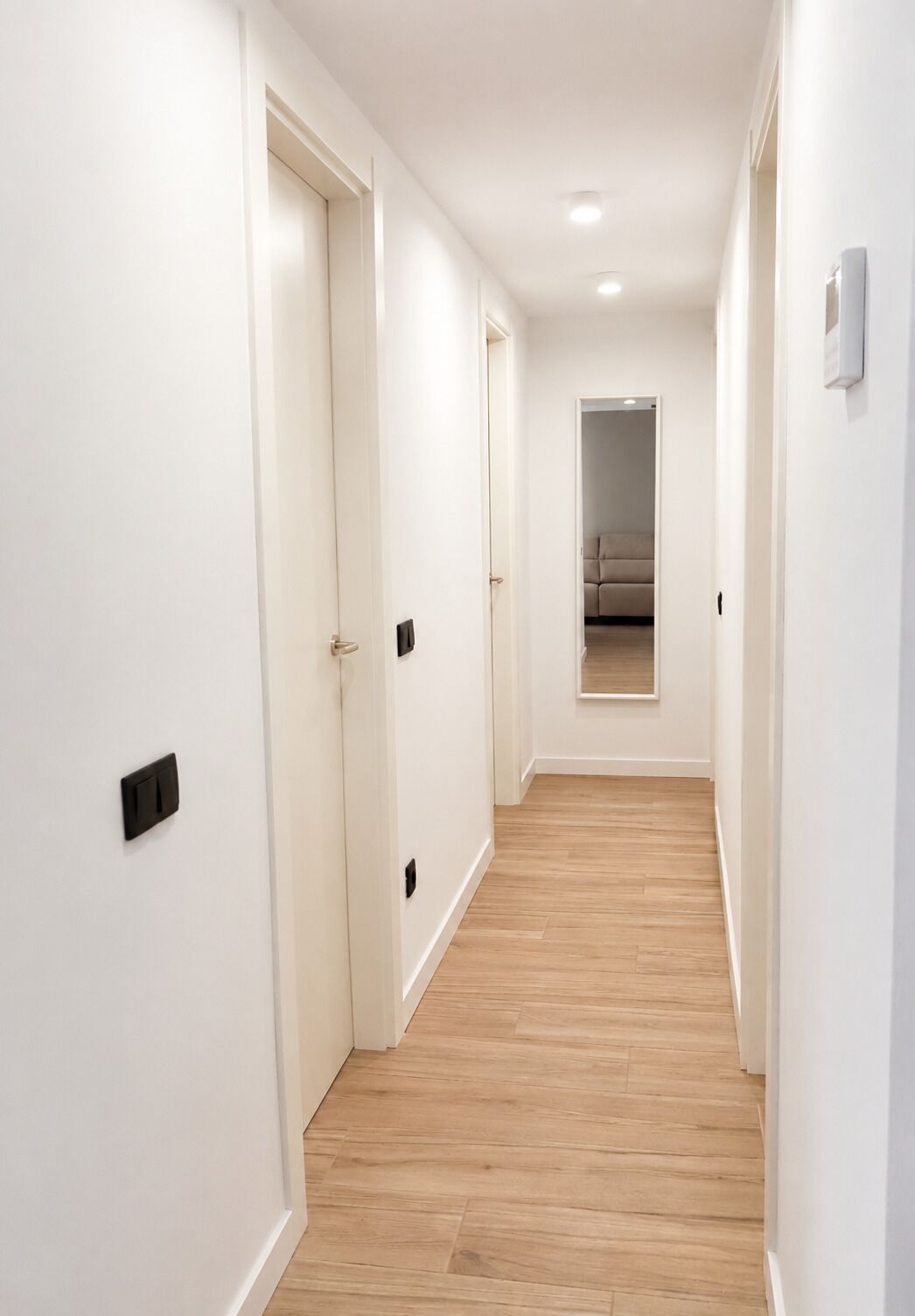 A hallway with white walls and wooden flooring, leading to a room with a beige sofa.