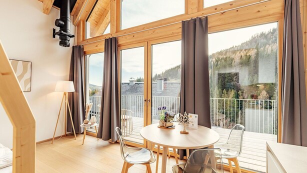 Tilava huone, jossa on suuri ikkuna, puiset kattopalkit, pyöreä ruokapöytä ja modernit tuolit.