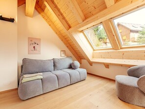 Ein Zimmer mit schrägen Wänden, Holzbalken, einem grauen Sofa, einem runden Sessel und einem Dachfenster mit Blick auf eine Wohngegend.