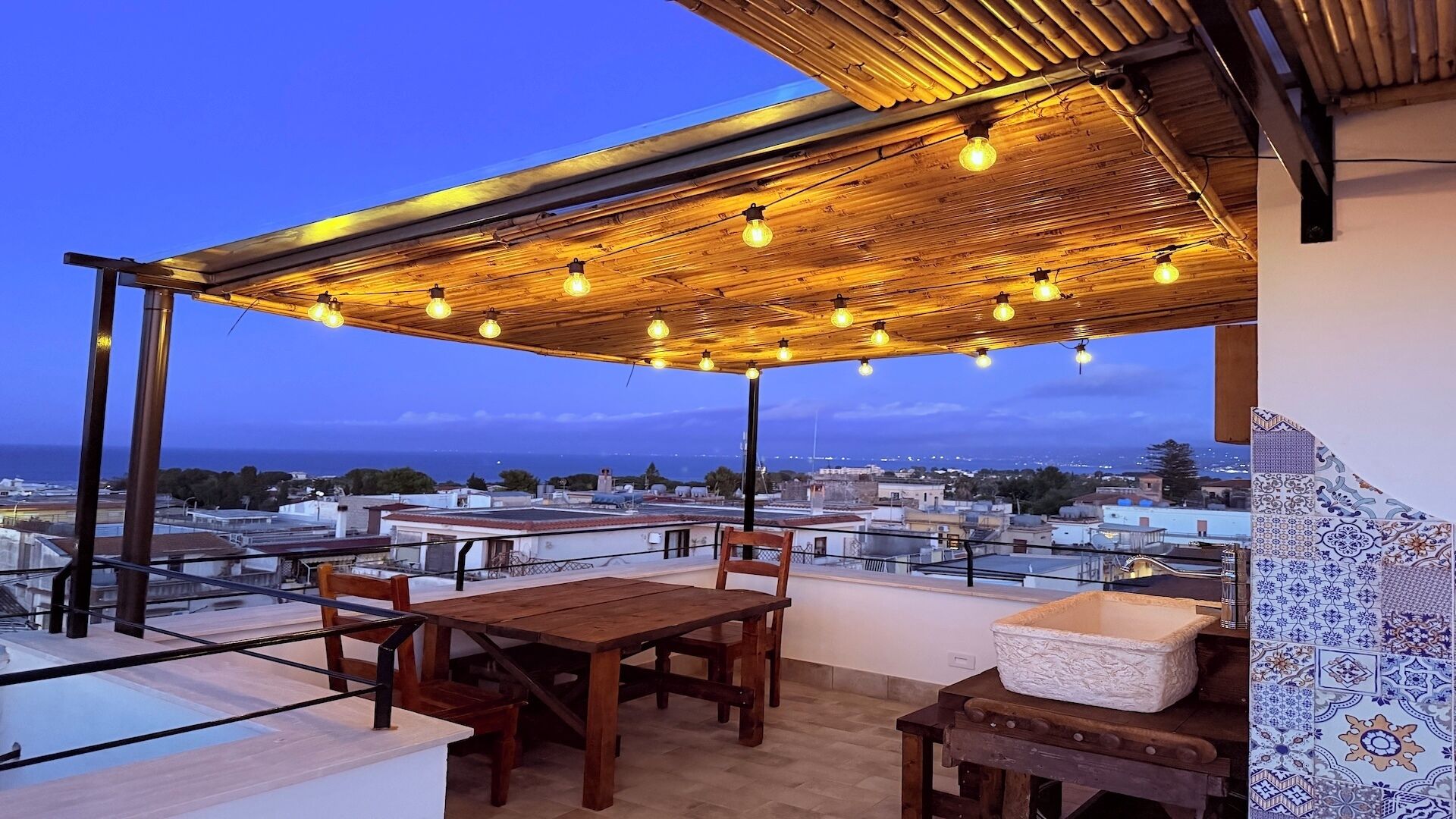 An outdoor terrace with a wooden table and chairs, a hanging light fixture, and a view of the cityscape.