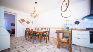A dining area with a wooden table and chairs, a chandelier, and a kitchen with white cabinets and a stove. - Solemar Sicilia - Stella Marina (Santa Flavia)