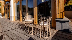 En träveranda med två fåtöljer i rotting och ett bord, med utsikt över en byggnad med stora fönster.