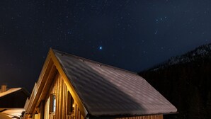 En stuga av trä med ett sluttande tak, ett fönster och en dörr, placerad mot en stjärnklar natthimmel.