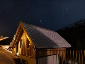 Eine Holzhütte mit geneigtem Dach, einem Fenster und einer Tür vor einem sternenübersäten Nachthimmel.