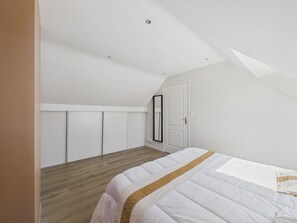 A bedroom with a sloped ceiling, a bed with a white and gold bedspread, a wardrobe, and a door. - Modern house for 7 with heated pool (Arradon)