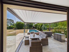 Une terrasse extérieure couverte avec une piscine, des meubles en osier et une vue sur le jardin arrière.