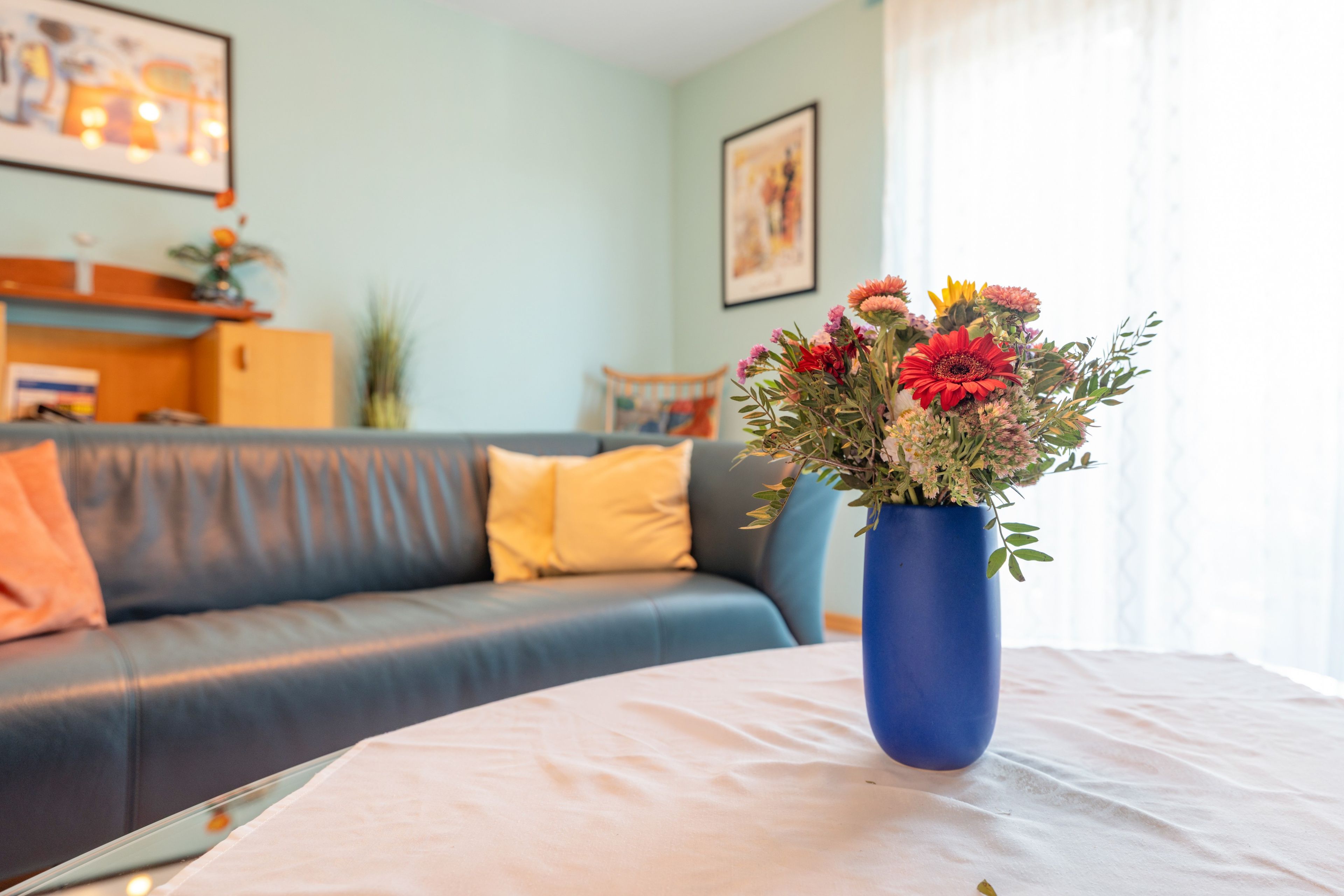 Ein Wohnzimmer mit einem blauen Sofa, einem weißen Tisch mit Blumenvase und einer hellblauen Wand mit einem gerahmten Bild.