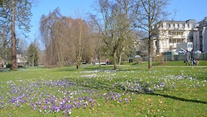 Um parque com área gramada e árvores, um edifício ao fundo e pessoas caminhando.