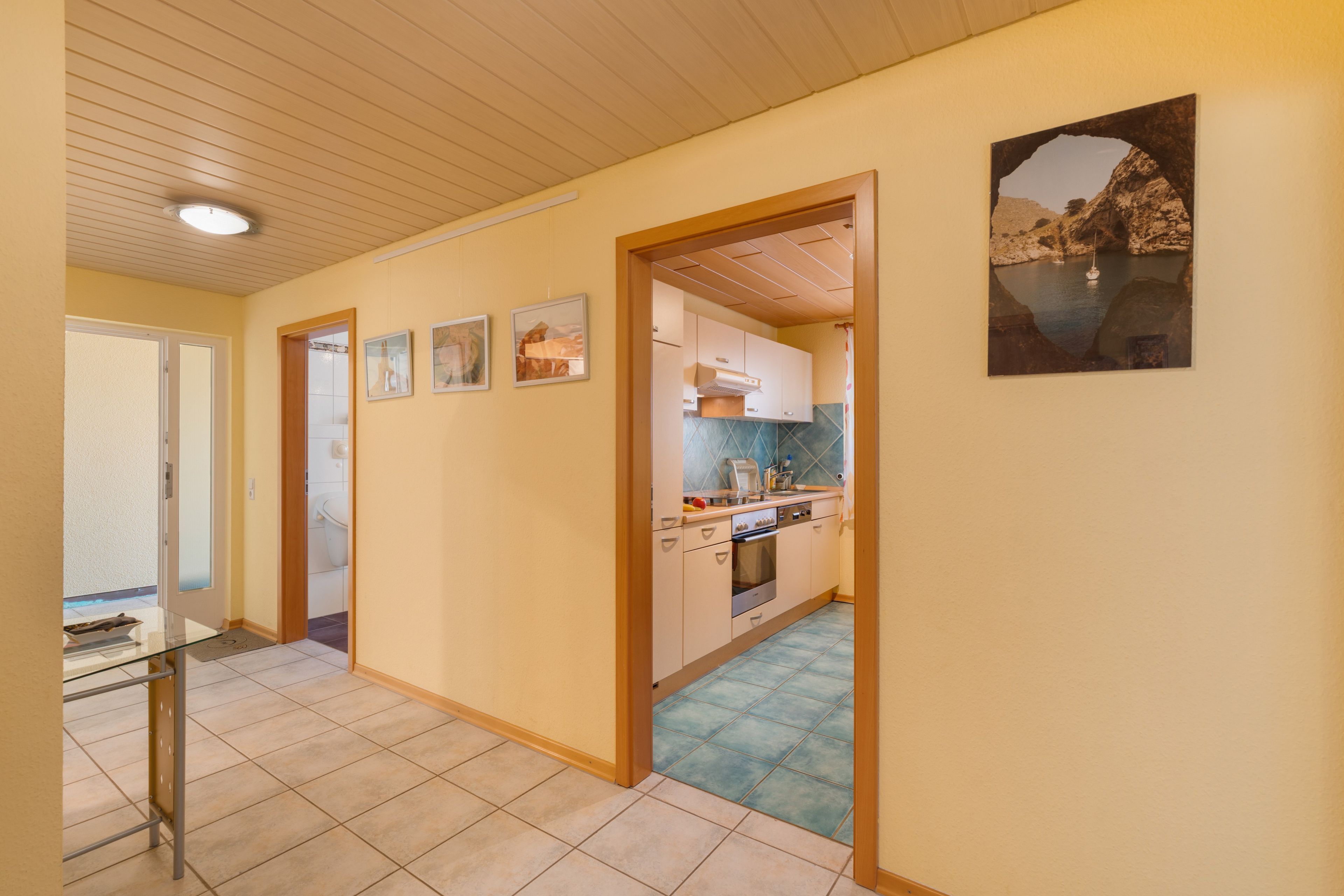 Ein Flur, der in eine Küche mit Fliesenboden und Holzbalkendecke führt, mit einem gerahmten Bild an der Wand.