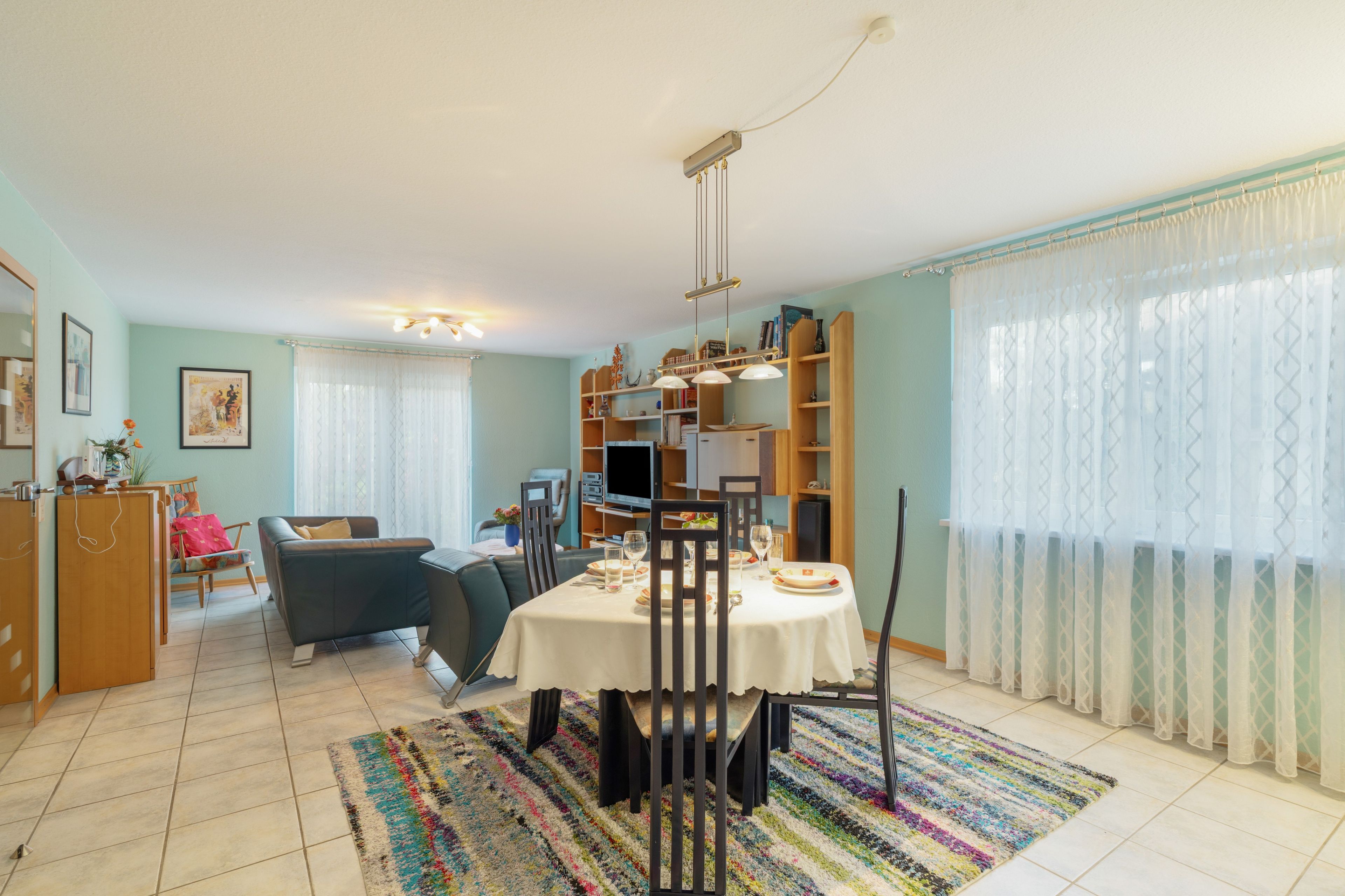 Uma sala de jantar com uma mesa posta para dois, um sofá e uma televisão ao fundo.