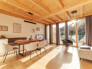 Ein modernes Wohnzimmer mit Holzboden, einem Essbereich mit Holztisch und -stühlen, einer Couch und einem Balkon mit Blick auf Bäume.
