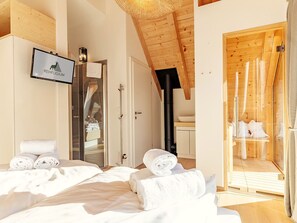 Ein modernes Schlafzimmer mit einem großen Bett, einem Fernseher, der 'REHFUGIUM' zeigt, einer Glasdusche und einem Saunabereich.