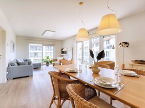 Una sala de estar moderna con un sofá, una mesa de comedor y sillas. Hay un televisor en la pared, una ventana con vistas y una planta en la mesa.