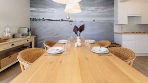 Ein Essbereich mit einem Holztisch, gedeckt für vier Personen, umgeben von Korbstühlen, einem großen Gemälde mit Strandmotiv und einer modernen Pendelleuchte.