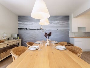 Ein Essbereich mit einem Holztisch, gedeckt für vier Personen, umgeben von Korbstühlen, einem großen Gemälde mit Strandmotiv und einer modernen Pendelleuchte.