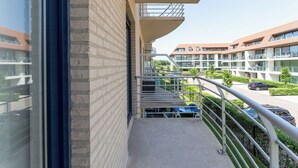Ein Balkon mit Metallgeländer, freie Sicht auf einen Parkplatz und ein Gebäude mit zahlreichen Fenstern und Balkonen.