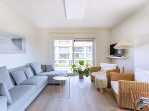 Una sala moderna con un sofá gris, una mesita redonda, una silla de mimbre, una lámpara de pie y un ventanal con vistas a edificios.