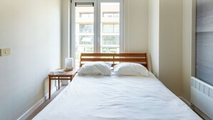 Ein Schlafzimmer mit einem Bett, einem Nachttisch, einer Lampe und einem Fenster mit Blick auf Gebäude.