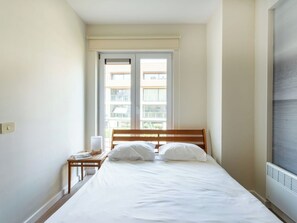 Ein Schlafzimmer mit einem Bett, einem Nachttisch, einer Lampe und einem Fenster mit Blick auf Gebäude.
