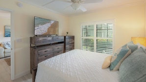 Une chambre à coucher comprenant un lit, une commode, un ventilateur de plafond, une fenêtre avec des stores et un tableau accroché au mur.