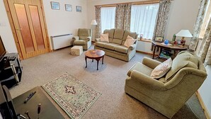 A living room with a sofa, armchairs, a coffee table, and a rug. There is a radiator, a door, and a window with curtains.