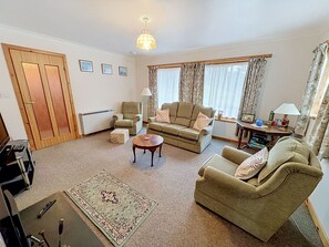 A living room with a sofa, armchairs, a coffee table, and a rug. There is a radiator, a door, and a window with curtains.