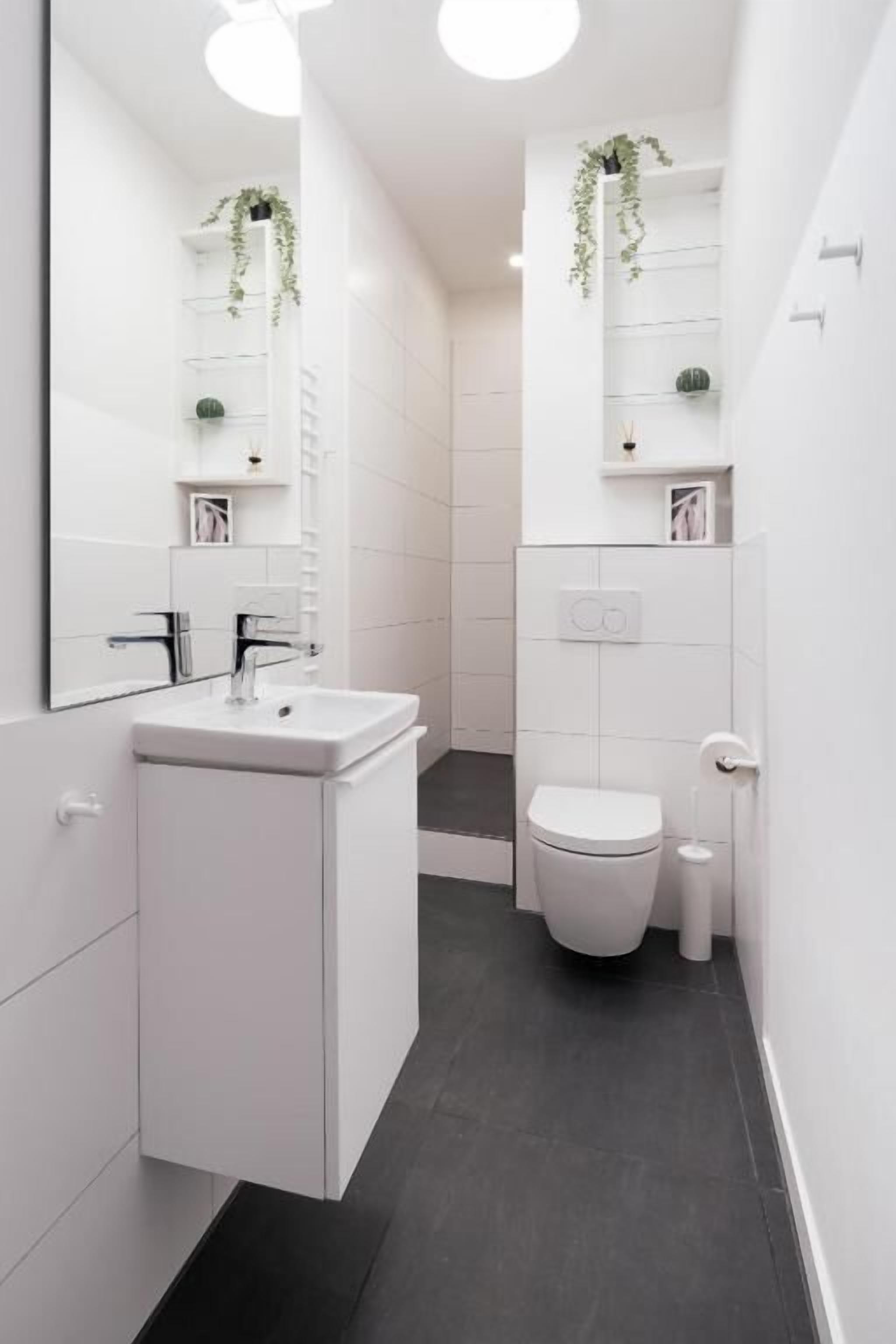 Une salle de bain moderne avec un lavabo, des toilettes et un miroir blancs, agrémentée d’un sol foncé et de murs blancs.