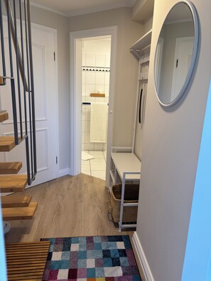 Un couloir avec un escalier en bois, une porte blanche, une salle de bain carrelée, une étagère blanche et un miroir.