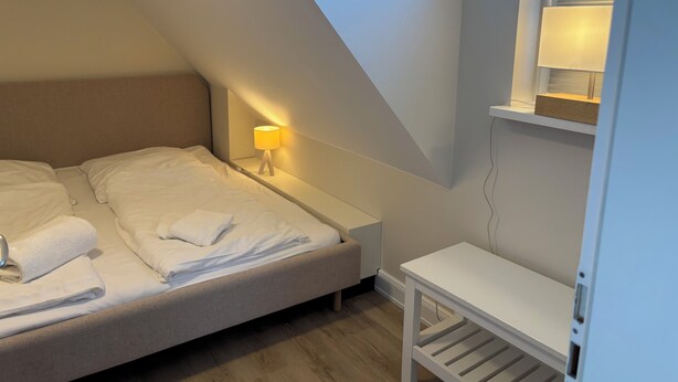 Een slaapkamer met een bed, nachtkastje, lamp en een klein wit nachtkastje.