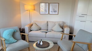 Een woonkamer met een bank, twee fauteuils, een salontafel, een vloerlamp en twee afbeeldingen in lijsten aan de muur.