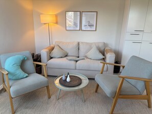 A living room with a sofa, two armchairs, a coffee table, a floor lamp, and two framed pictures on the wall. - 26 West – Duplex for 4, Westerland, 2 bathrooms, west-facing balcony, 10 min. to the beach (Sylt)