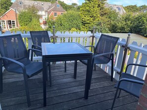 A deck with a table and chairs, surrounded by a white picket fence, with houses and trees in the background. - 26 West – Duplex for 4, Westerland, 2 bathrooms, west-facing balcony, 10 min. to the beach (Sylt)