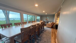 Een eetgedeelte met een lange tafel en stoelen, een bank, en uitzicht op een grasveld door grote ramen.