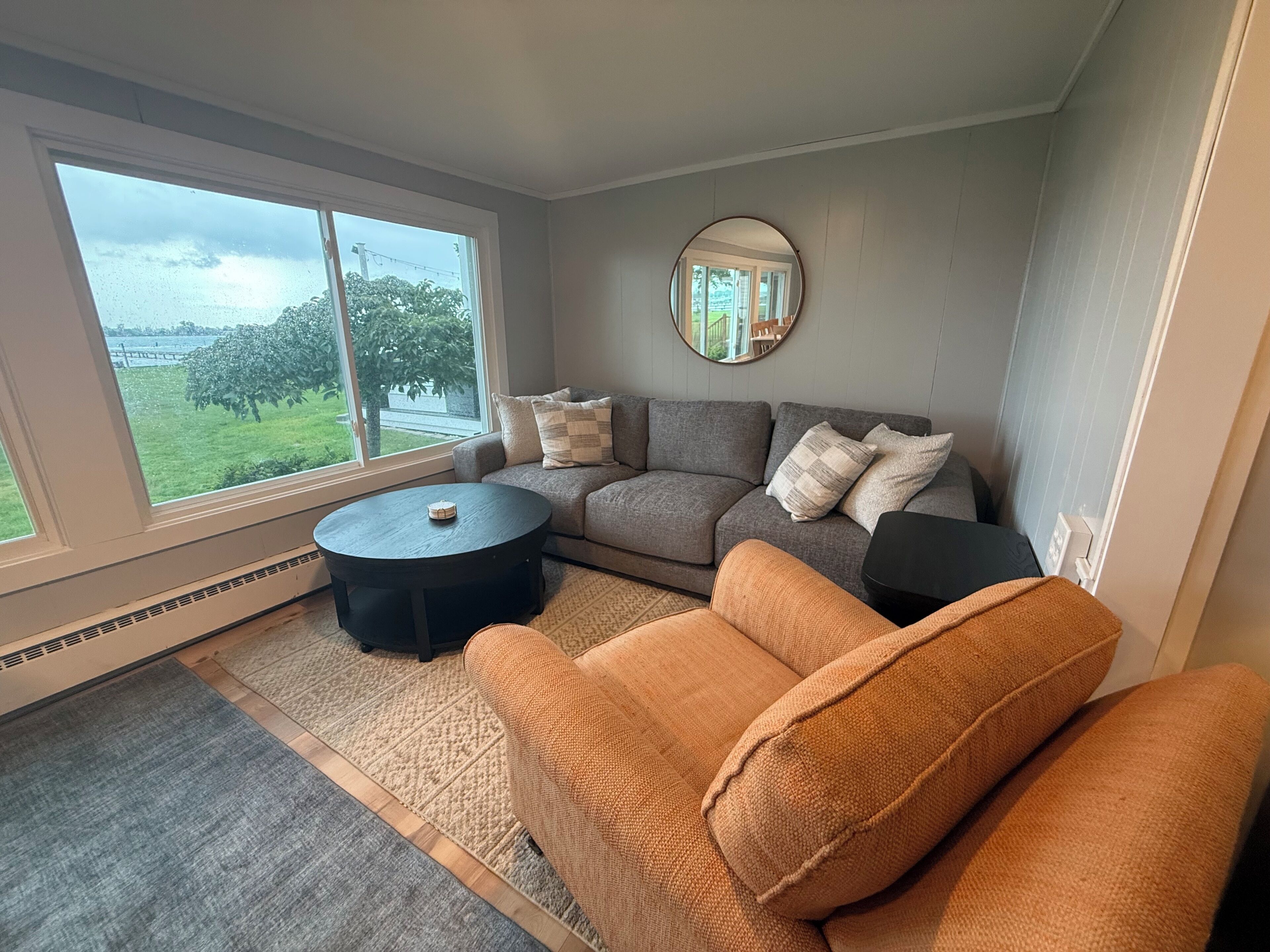 A living room with a gray sofa, a round coffee table, and a view of a green landscape through large windows.