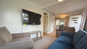 Een woonkamer met een flatscreen televisie aan de muur, een grijze bank, een blauwe bank en een houten salontafel.