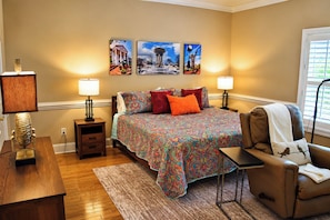 A bedroom with a bed, two bedside lamps, a nightstand, a chair, a window with blinds, and wall art.