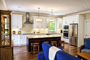 A modern kitchen with white cabinets, a central island with stools, a stainless steel refrigerator, and a large window with greenery. - The Cary Retreat • Luxury Cottage in the Heart of Downtown Cary (Cary)