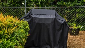 Een barbecue bedekt met een zwarte doek, omringd door groen en een pad van stenen.