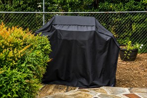 A barbecue grill covered with a black cloth, surrounded by greenery and a stone pathway.