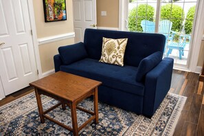 A blue sofa with a patterned pillow, a wooden coffee table, and a patterned rug in a room with a door and a window with a view of greenery.