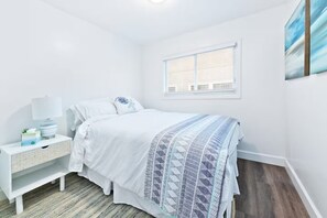 Ein Schlafzimmer mit Bett, Nachttisch, Fenster und einem an der Wand hängenden Kunstwerk.